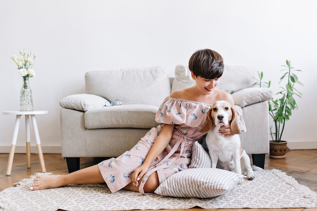 Free Photo glad young woman with shiny brown hair posing on the floor with her cute beagle puppy. indoor portrait of excited girl in dress with floral print sitting on the carpet with dog