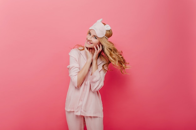 Glamorous curly girl expressing energy in morning. Pleasant caucasian woman in silk pyjama posing on pink wall.