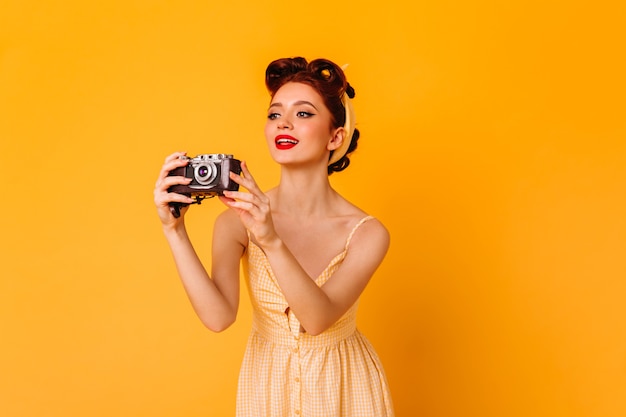 Glamorous pinup girl taking pictures. Inspired ginger woman with camera standing on yellow space.