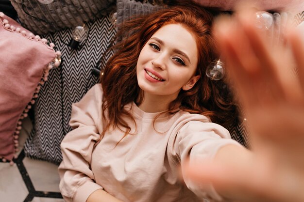 Glamorous redhaired girl lying on the floor Charming relaxed woman making selfie