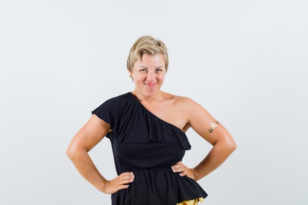 Glamorous woman in black blouse standing with hands on waist and looking jolly 