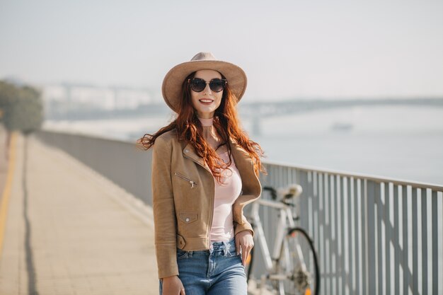 Glamorous woman in black sunglasses posing on bridge with bicycle on wall