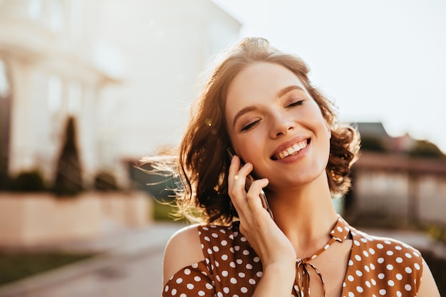 Glamorous young woman talking on phone with eyes closed. Outdoor shot of pretty caucasian girl with short brown hair.