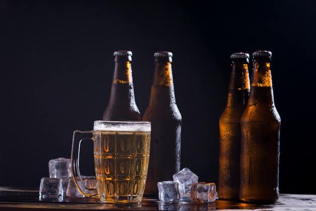 Glass bottles of beer with glass and ice on dark background