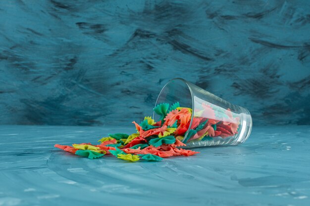 Glass of colorful raw farfalle pasta on blue background. 