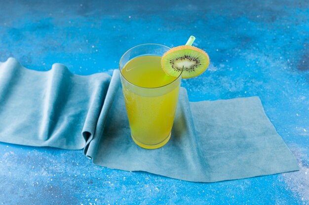 Glass cup of fresh juice with slice of kiwi and straw placed on tablecloth .