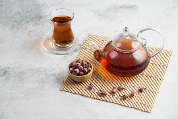 A glass cup of tea with dried roses and teapot