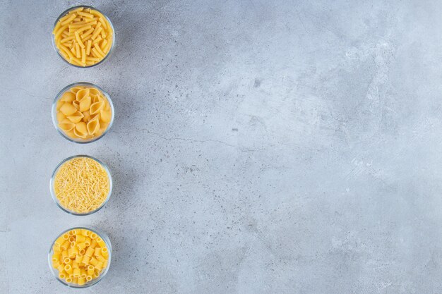 Glass cups full with different types of raw dry pasta on a stone background.