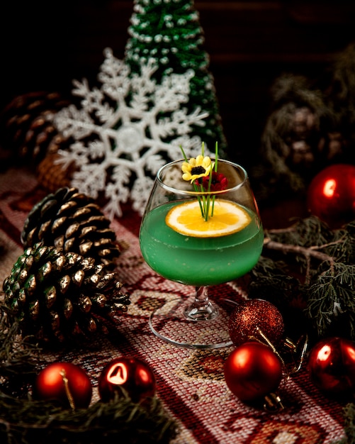 A glass of green drink garnished with orange slice and fake flowers around christmas decorations