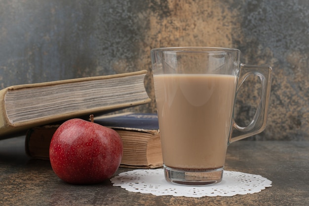 A glass of hot coffee with one red apple and books on marble wall.