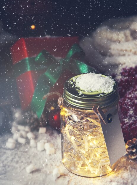 Glass jar with lights with a gift next to it while it snows viewed from above