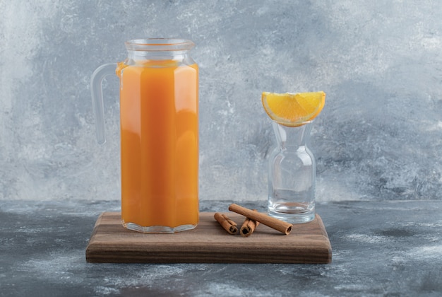 Glass jug of fresh juice and cinnamon sticks on wooden board. 