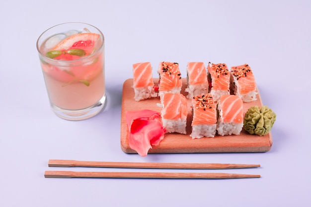 A glass of juice with classic salmon sushi; wasabi and pickled ginger on chopping board with chopsticks against white backdrop