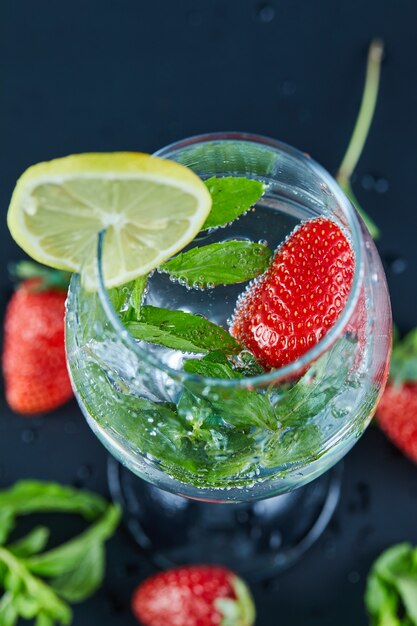 A glass of juice with whole fruits and slice of lemon inside on dark surface