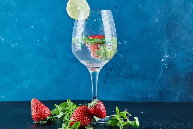 A glass of juice with whole fruits and slice of lemon inside on dark surface