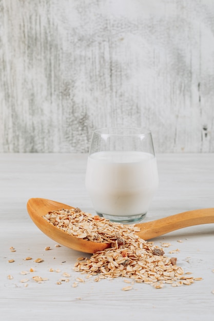 Glass of milk with oats in wooden spoon side view on a white wooden background