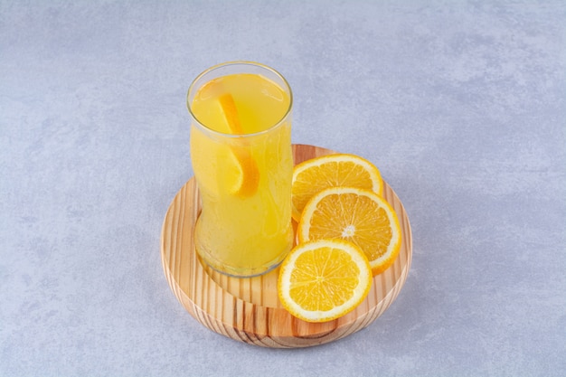 A glass of orange juice next to slice orange on a wooden plate on marble table.