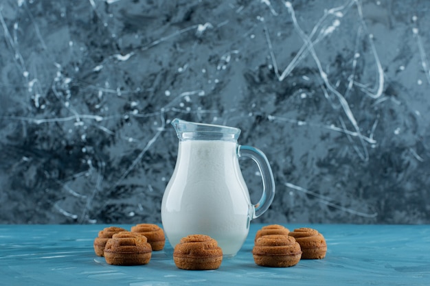 Free photo a glass pitcher of fresh milk with sweet round cookies on a blue surface