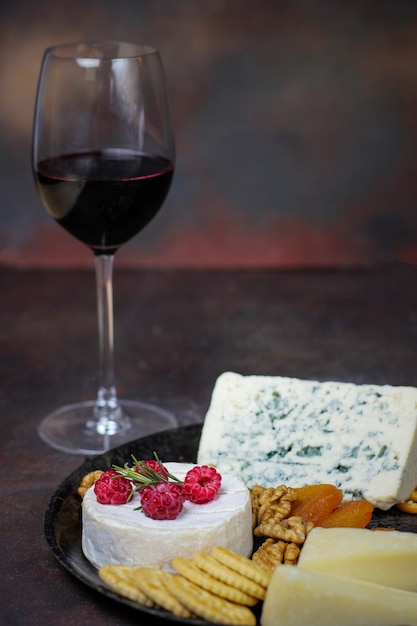 Glass of red wine with cheese plate on dark  with camembert cheese,blue cheese,gauda and berries and snacks