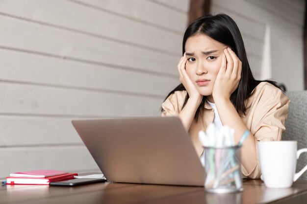 Gloomy asian female student sitting bored with laptop at home tired or bored of studying having onli...