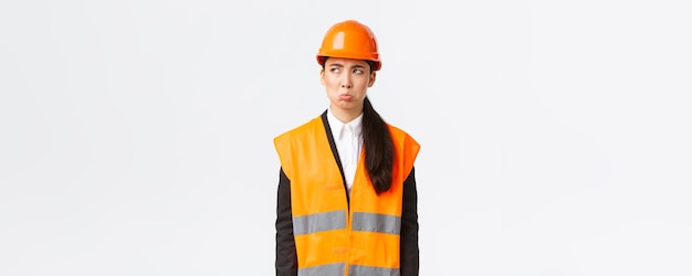 Gloomy and offended young asian female construction manager industrial woman in safety helmet complaining or whining pouting sad and looking upper left corner disappointed