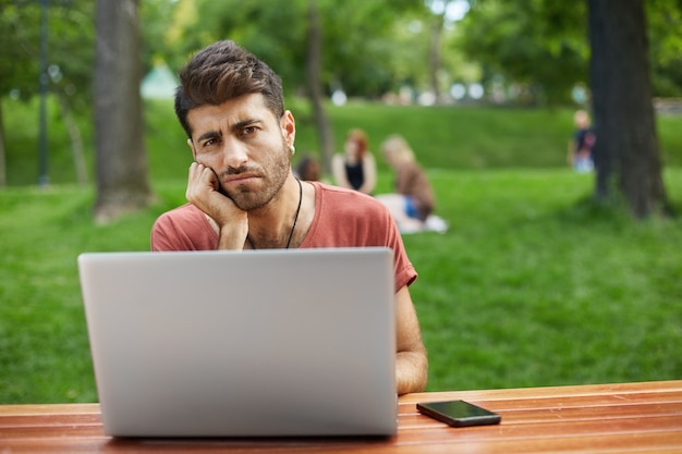 Gloomy and sad guy sitting in park with laptop, looking upset