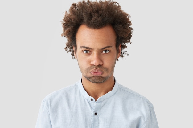 Gloomy sullen man with curly hair, has stubble, displeased expression, expresses negative emotions, dressed in white shirt