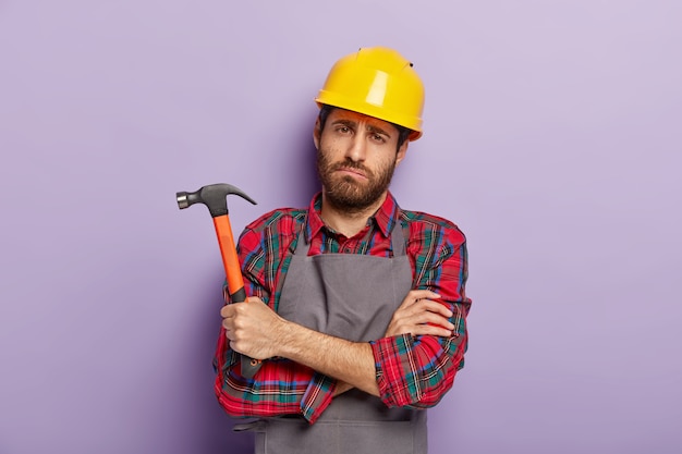 Free photo gloomy unhappy male repairer has sad tired look, keeps hands crossed, holds hammer in hand, fatigue after repairing and manual work, wears special uniform. handcrafting, hammering, building.