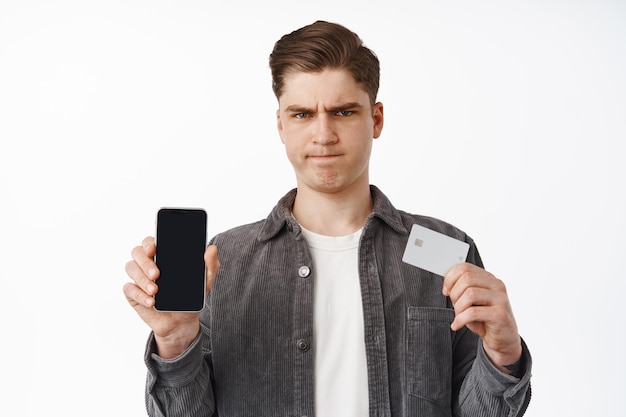 Gloomy and upset guy showing credit card with smartphone screen, frowning angry, disappointed with online shopping, finance app, white background. Technology concept