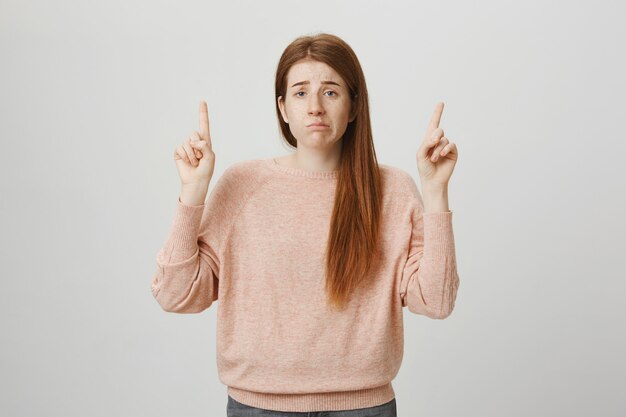 Gloomy upset redhead girl pouting indecisive, pointing fingers up