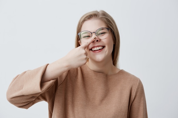 Gloomy young caucasian female makes fun, touches her nose, mocks, laughs and broadly smiles, demonstrates her white even teeth. Positive blonde girl joyfully smiles 