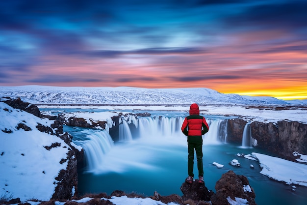 Free photo godafoss waterfall at sunset in winter, iceland. guy in red jacket looks at godafoss waterfall.