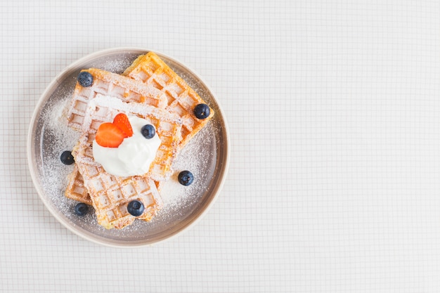 Free photo golden brown waffle topped with sliced strawberries; blueberries and whipped cream on plate