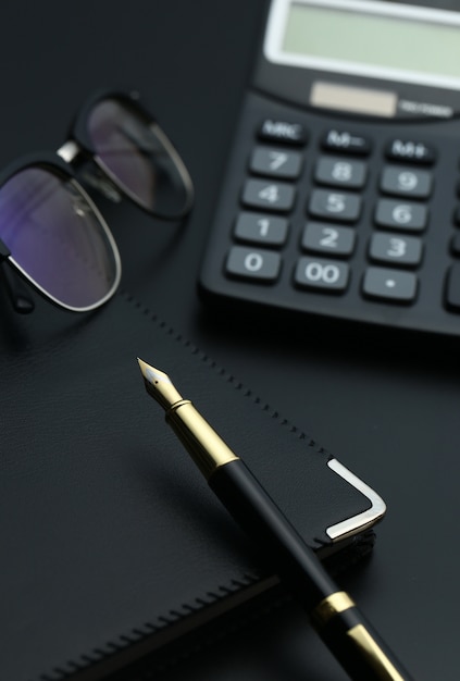 Free photo golden pen,notebook,calculator and glasses on black desk