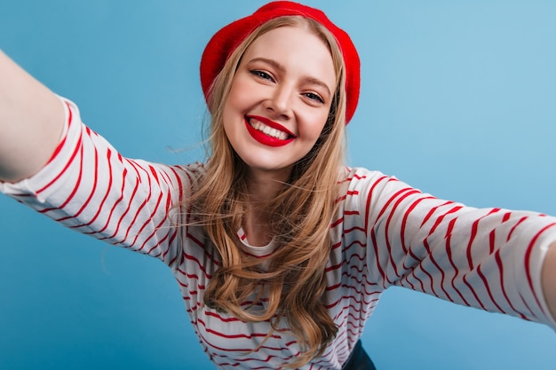 Free photo good-humoured french girl taking selfie with smile. happy blonde female model posing on blue wall.