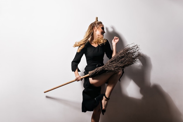 Free Photo good-humoured girl in black outfit dancing with broom in halloween. indoor photo of glad blonde lady in carnival costume expressing positive emotions.