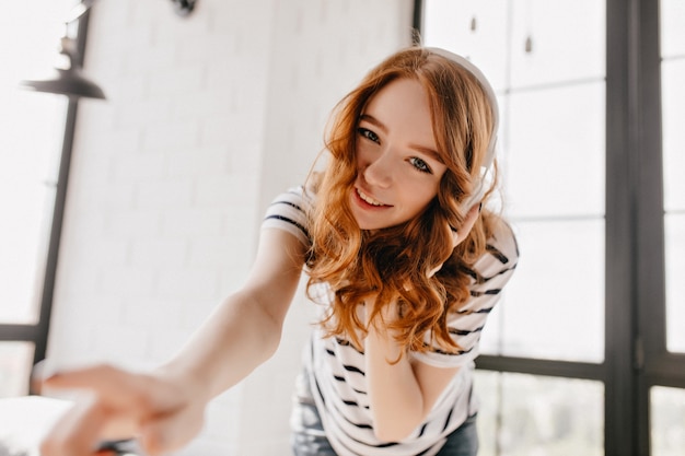 Good-humoured girl in headphones looking. Charming curly female model having fun at home.