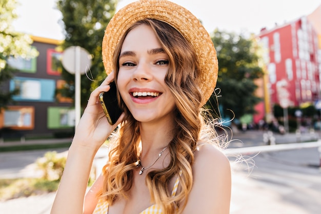 Good-humoured white girl talking on phone in summer day. Outdoor portrait of spectacular lady in hat holding smartphone on street.