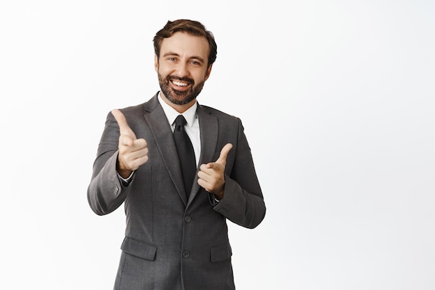 Free photo good job smiling boss pointing fingers at camera showing thumbs up looking pleased at employee wearing suit white background