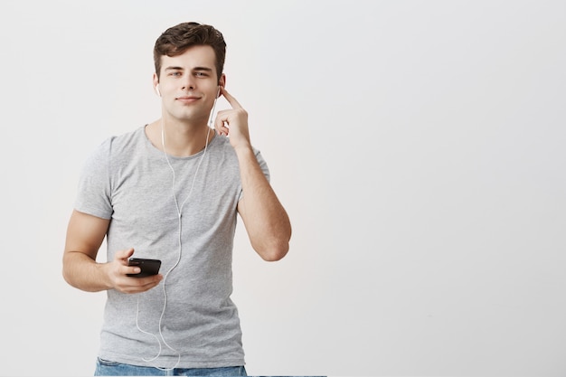 Good-looking attractive youngman in gray t-shirt having pleased look,holding mobile phone in his hand, wearing white earphones on, joyfully listens to his favourite songs, using music app.