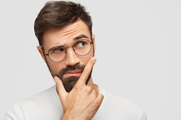 good looking bearded young man holds chin and looks pensively aside, raises eyebrows and ponders about something