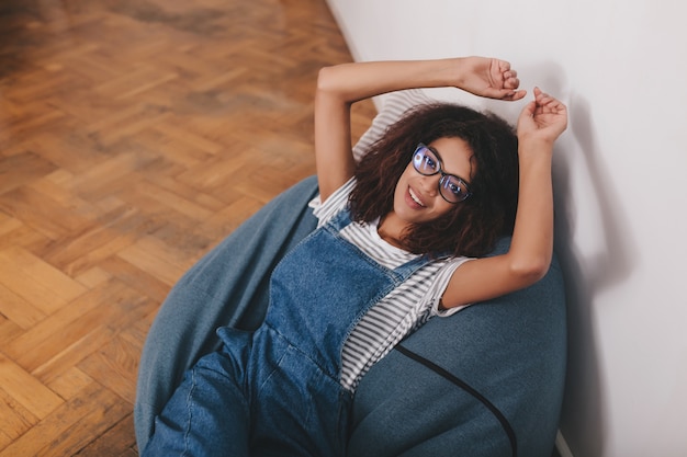 Good-looking black girl wears striped shirt and denim overall resting at home lying on pillow with hands up