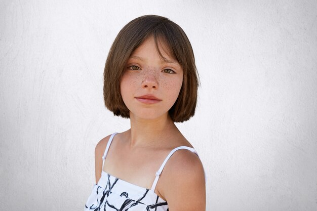 Good-looking brunette kid with freckles and short hair posing against white concrete wall dressed in white dress. Little child with dark wide eyes and freckled skin isolated over white wall