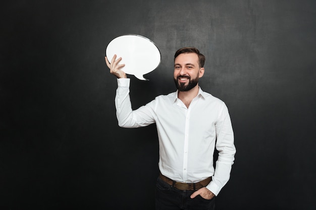 Free Photo good-looking brunette man holding blank speech bubble and looking at camera over dark gray wall copy space