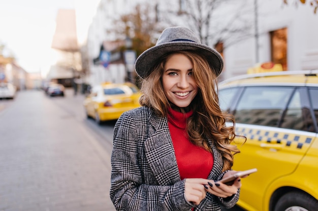 Free photo good-looking businesslady in gray hat gently smiling, walking down the road. stunning dark-haired female model in coat standing beside taxi in morning.