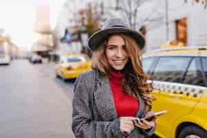 Free photo good-looking businesslady in gray hat gently smiling, walking down the road. stunning dark-haired female model in coat standing beside taxi in morning.