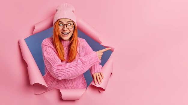 Good looking cheerful redhead woman pointing at copy space, wears casual knitted sweater and hat.