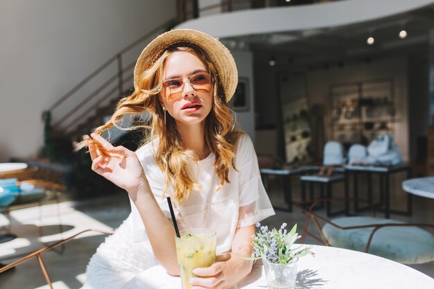 Free Photo good-looking girl playing with her curly hair resting at the table with little flower vase on it