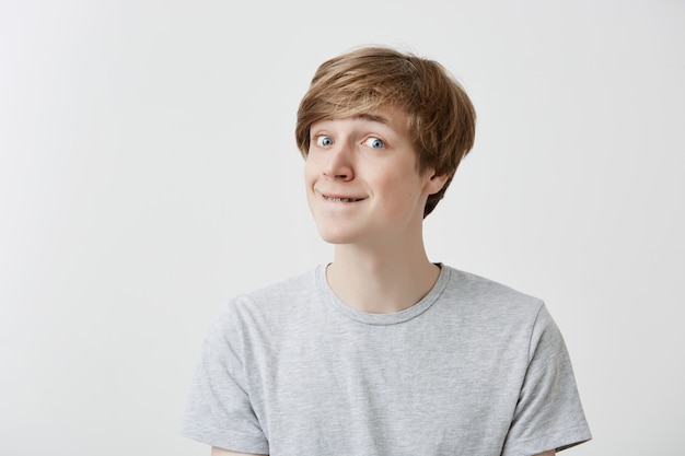 Good-looking male model with braces presses lips, looks pensively aside, thinks over his future actions or plans, wears light gray t-shirt, has clever expression, isolated against gray background