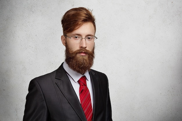 Free photo good-looking stylish redhead entrepreneur or office worker with thick beard and rectangle glasses posing isolated against gray wall  with copy space for your text or promotional content